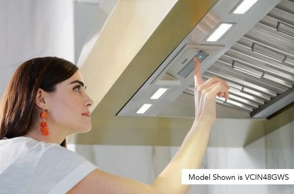 Woman adjusting controls on stainless steel custom insert range hood in modern kitchen setting