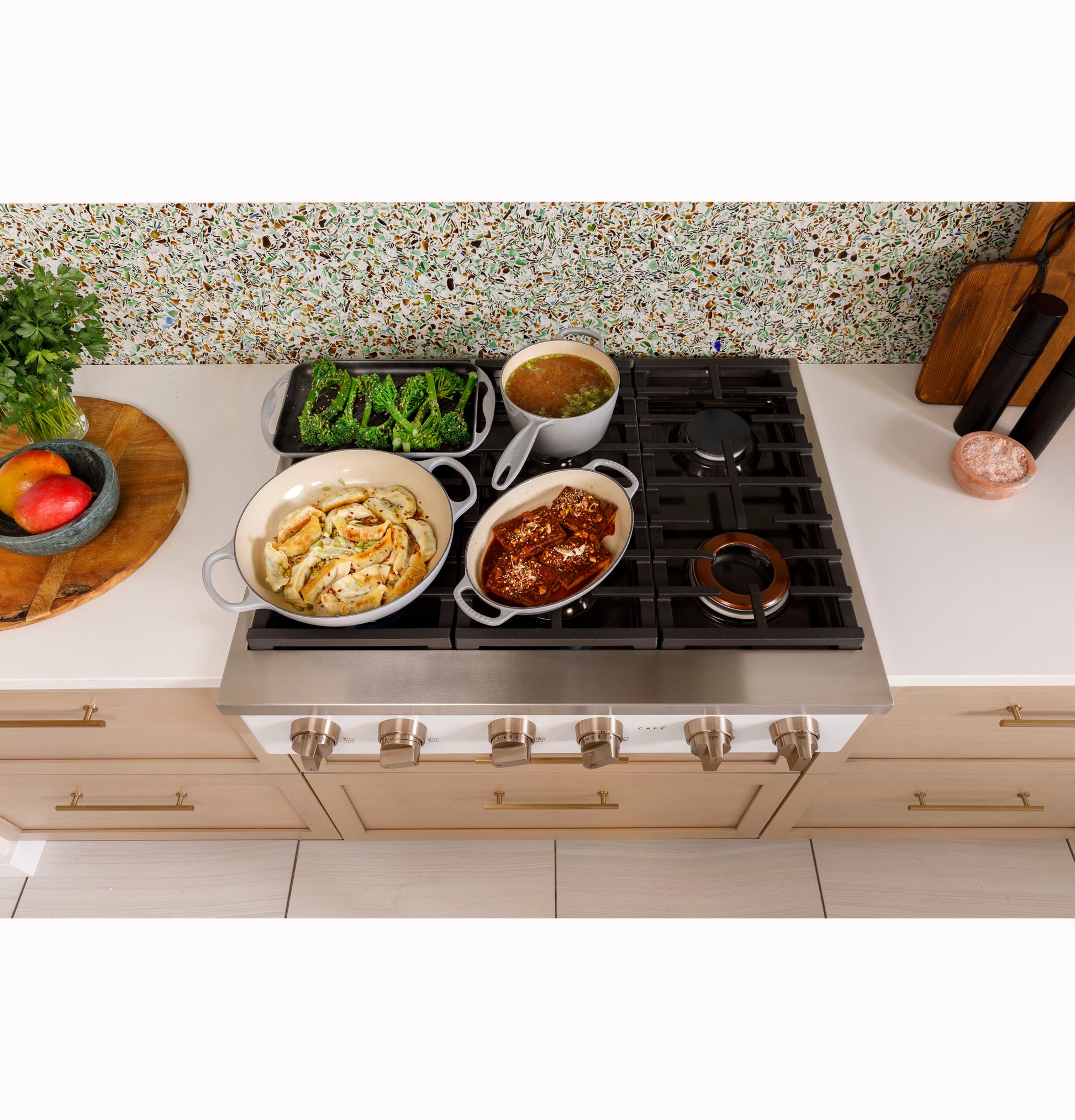 Commercial-style stainless steel gas rangetop with six burners cooking diverse dishes in bright kitchen setting