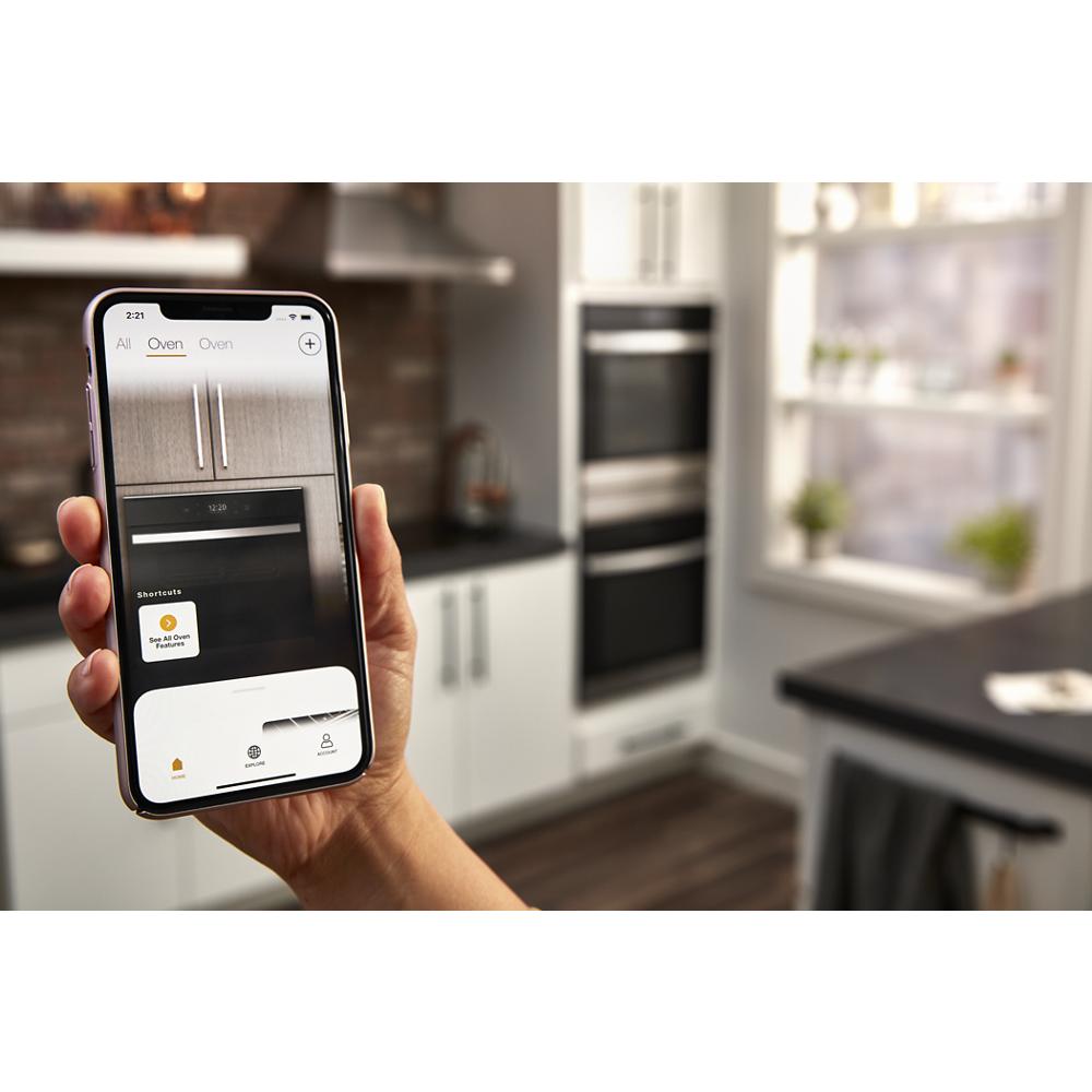 Hand holding smartphone displaying smart oven control app with double wall ovens in bright kitchen setting