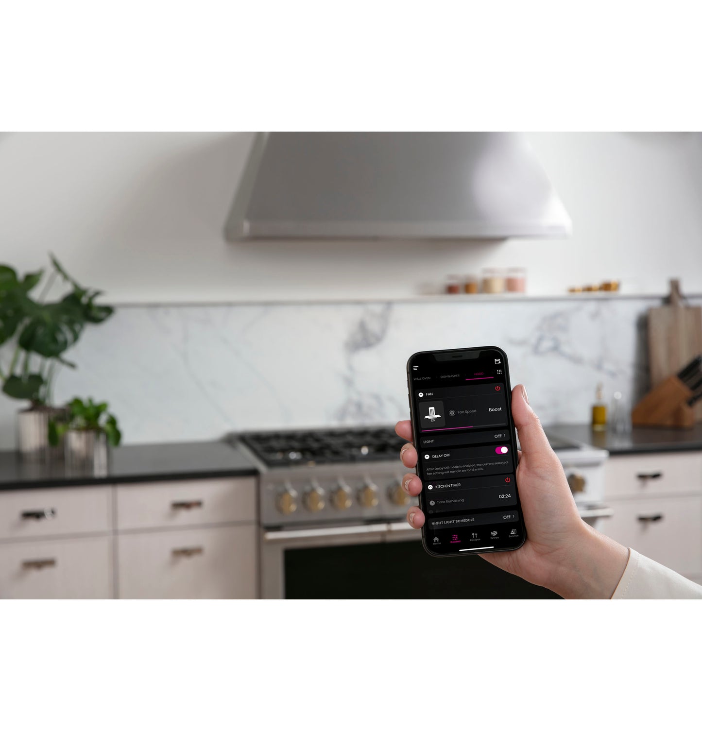Hand holding smartphone displaying app in modern kitchen with stainless steel professional range hood and gas stove
