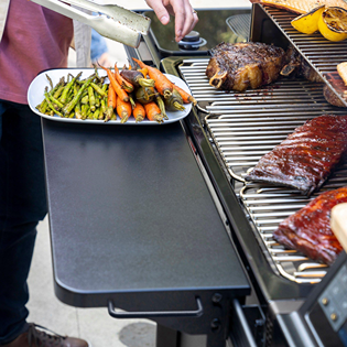Matte black folding front shelf attached to grill with cooked meat and vegetables for outdoor cooking and grilling tasks