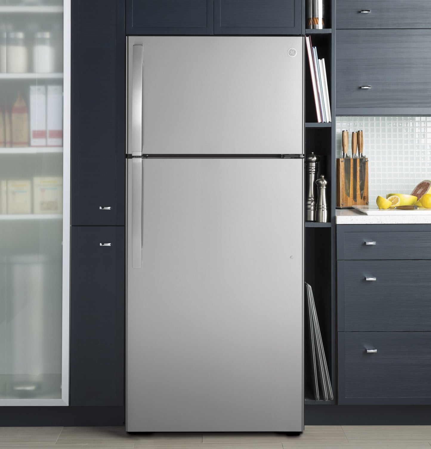 Stainless steel top-freezer refrigerator integrated into dark blue kitchen cabinetry with countertop and cutting board nearby
