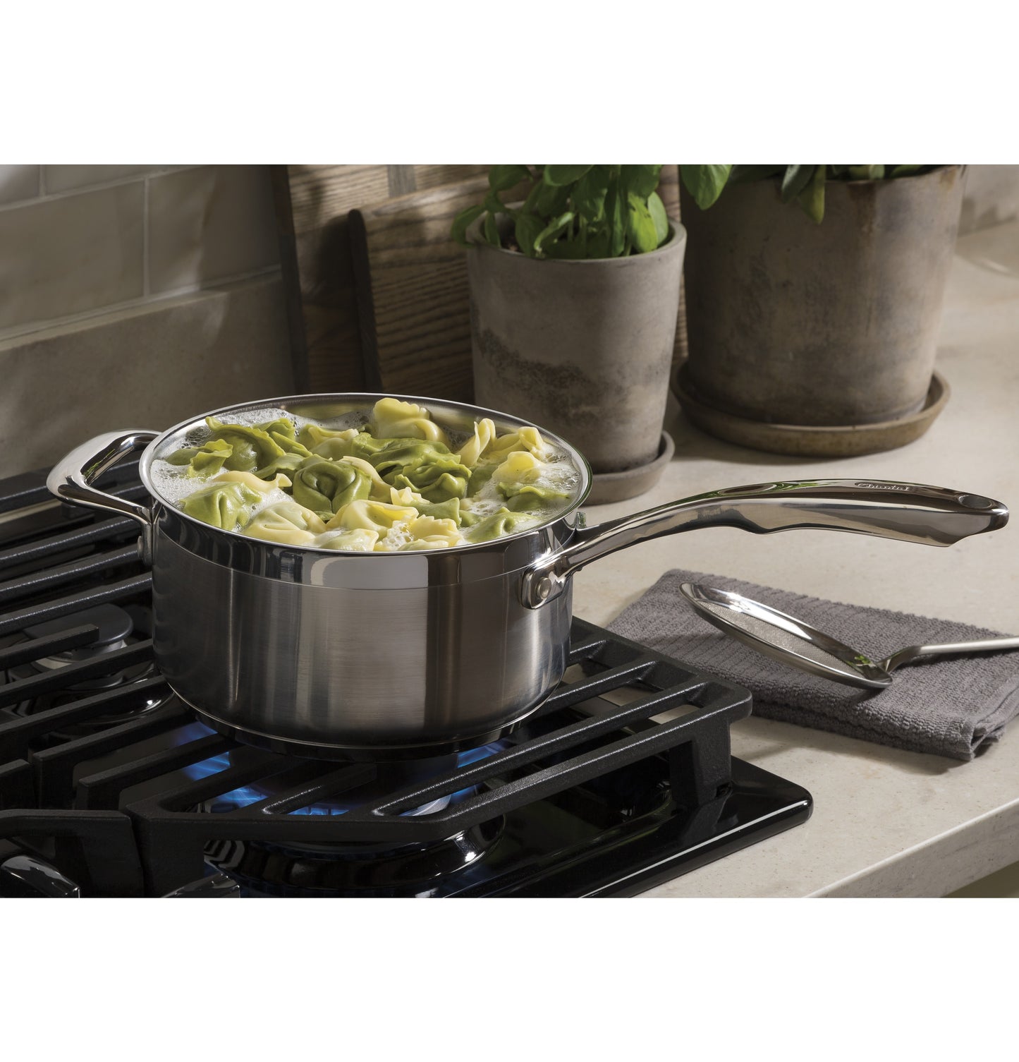 Stainless steel pot with boiling pasta on a gas burner cooktop surrounded by plants and kitchenware tools.