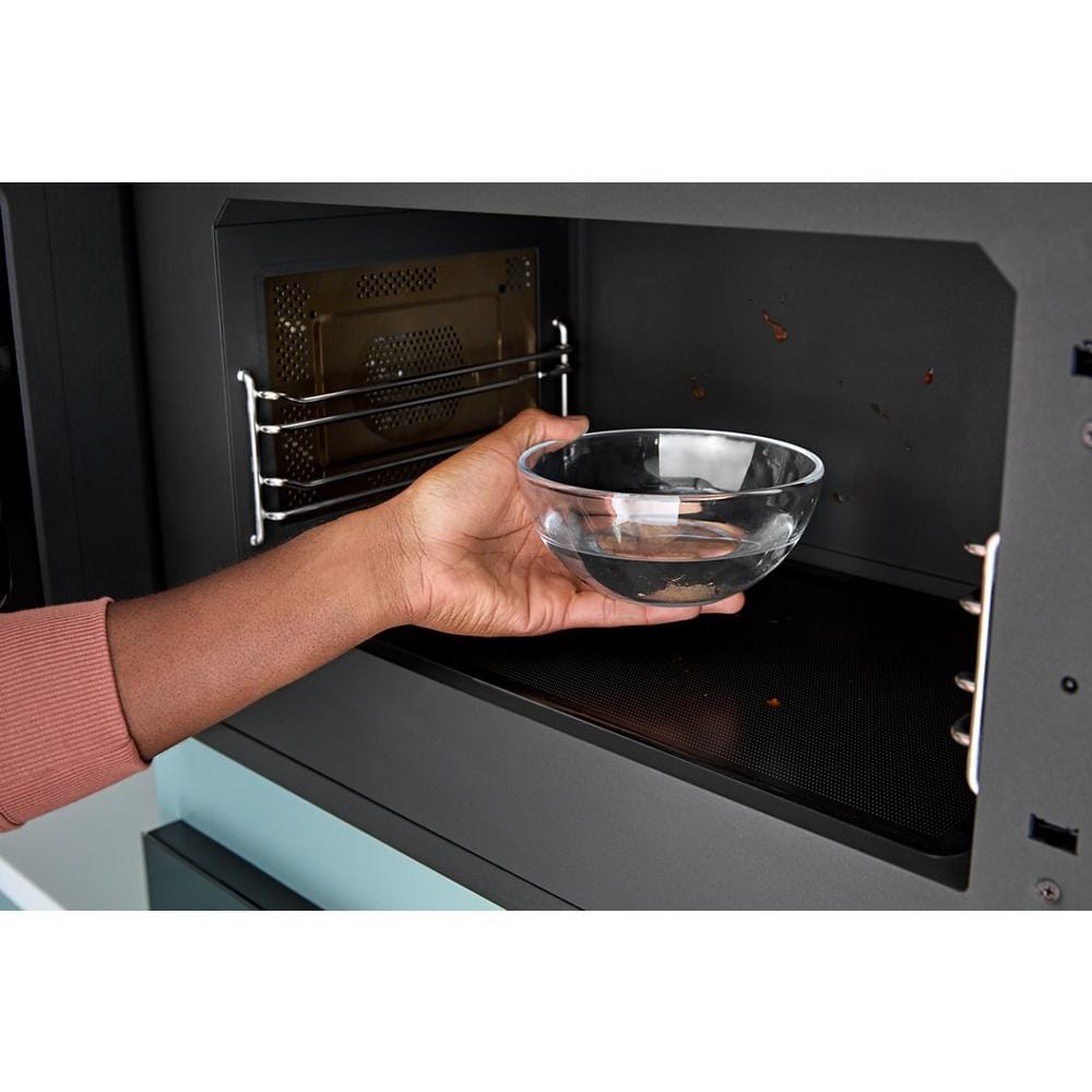 Hand placing clear glass bowl inside built-in multifunctional microwave oven with black interior for kitchen appliances.