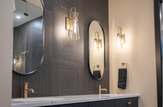 Modern bathroom vanity featuring a white marble countertop, dual brushed brass faucets, oval mirrors, and wall-mounted glass light fixtures against a textured dark accent wall.