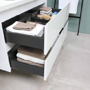 Modern white bathroom vanity with two open drawers containing neatly folded towels, bath accessories, and personal care items on a light tile floor.