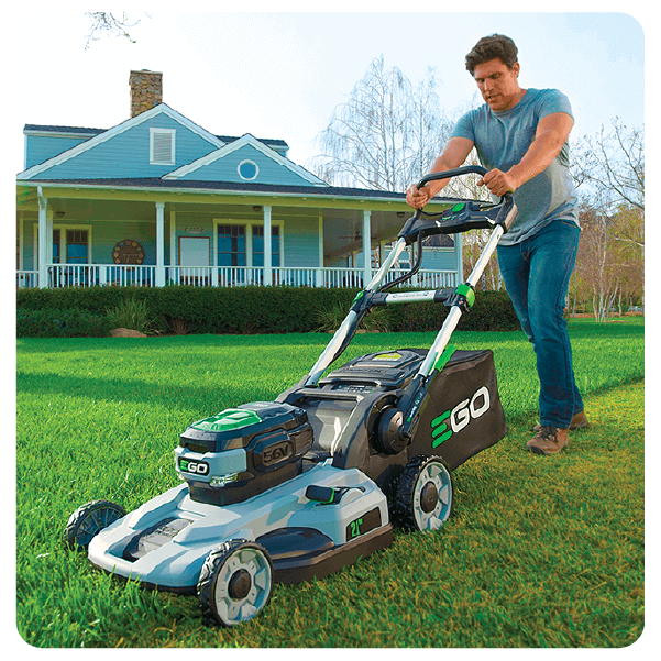 Man wearing a blue t-shirt and jeans pushing a cordless electric lawn mower on a green lawn in front of a suburban house during daytime.