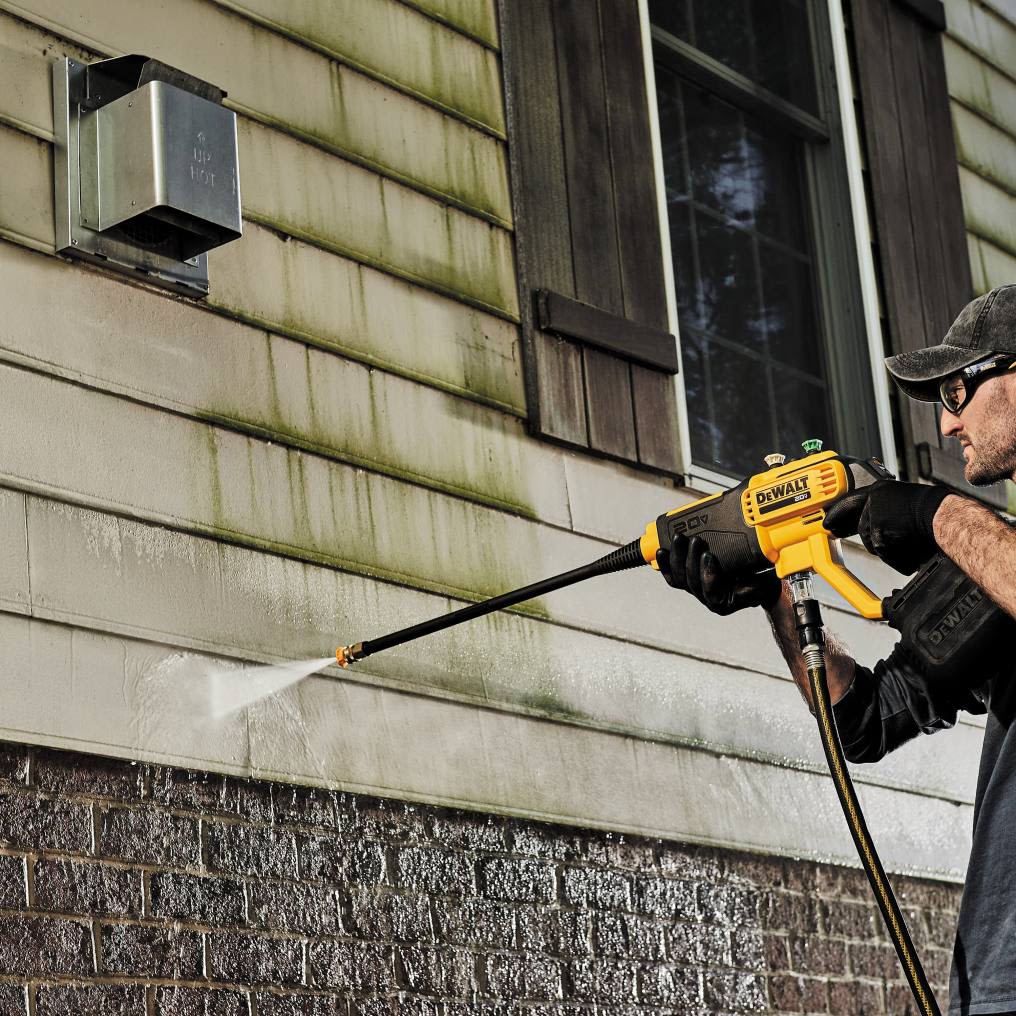 Man using yellow and black cordless power cleaner to remove dirt and grime from building exterior siding and brick wall
