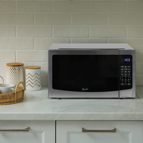 Compact stainless steel microwave oven with digital display on white kitchen countertop near mugs and storage jars