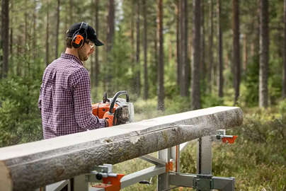 Man wearing protective gear operating a portable chainsaw mill for outdoor timber cutting in a forest setting