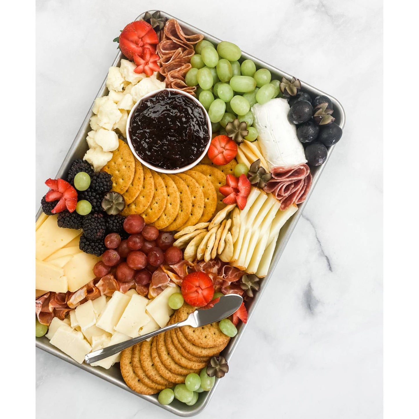 Metal pan filled with assorted cheeses, crackers, grapes, blackberries, meats, and a small bowl of jam on marble surface
