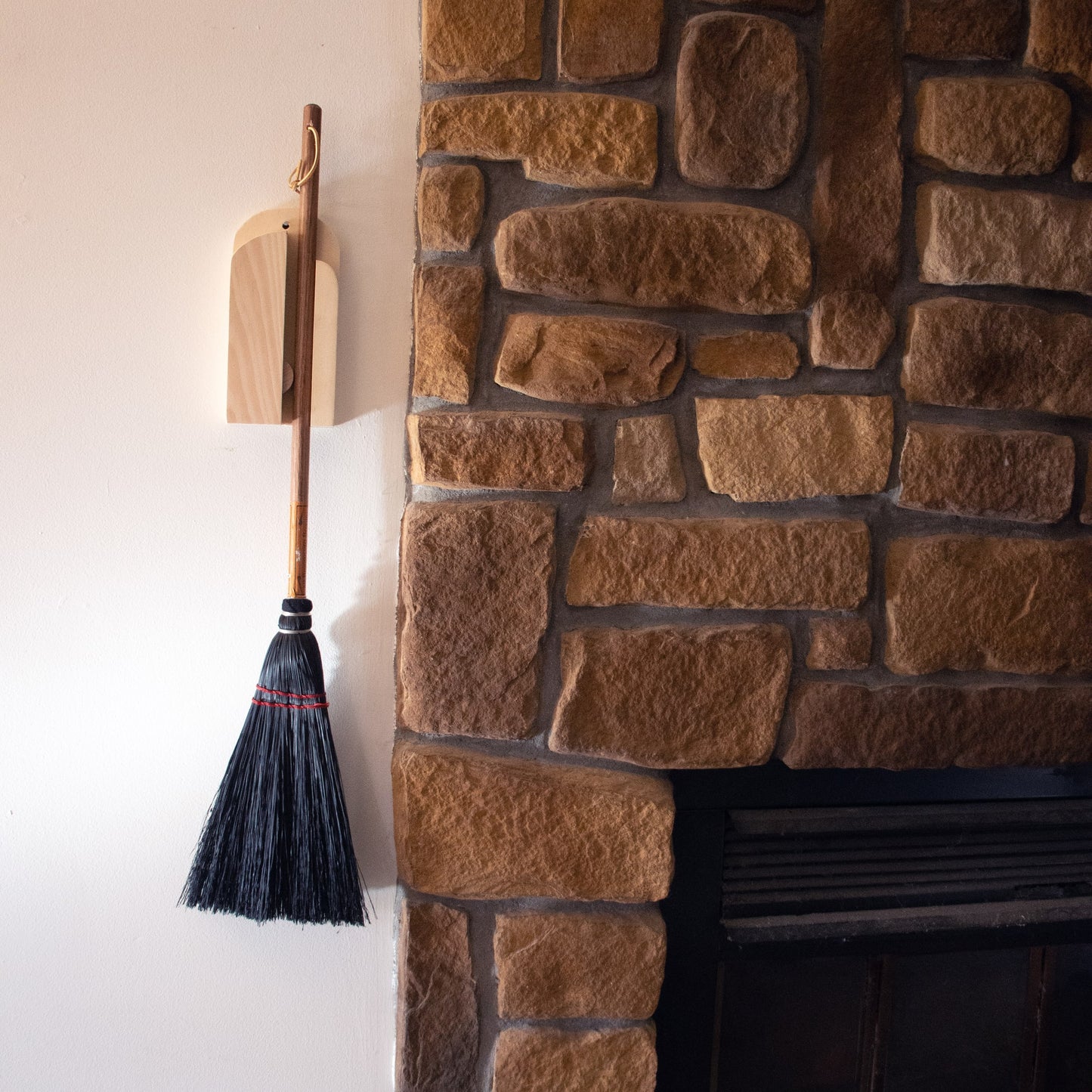 Wooden wall-mounted broom holder with a natural finish holding a broom beside a rustic stone fireplace in a home improvement setting