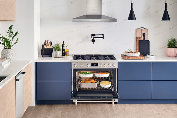 Kitchen scene with stainless steel range, open oven with cooked dishes, modern wall-mounted vent hood, and blue cabinetry