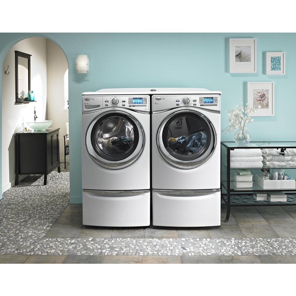 Pair of white front load washer and dryer on storage pedestals in laundry room with teal walls and decorative shelving