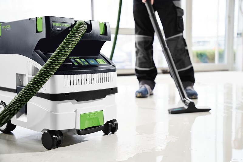 Portable Festool dust extractor with green hose in a bright workshop cleaning liquid from floor surface