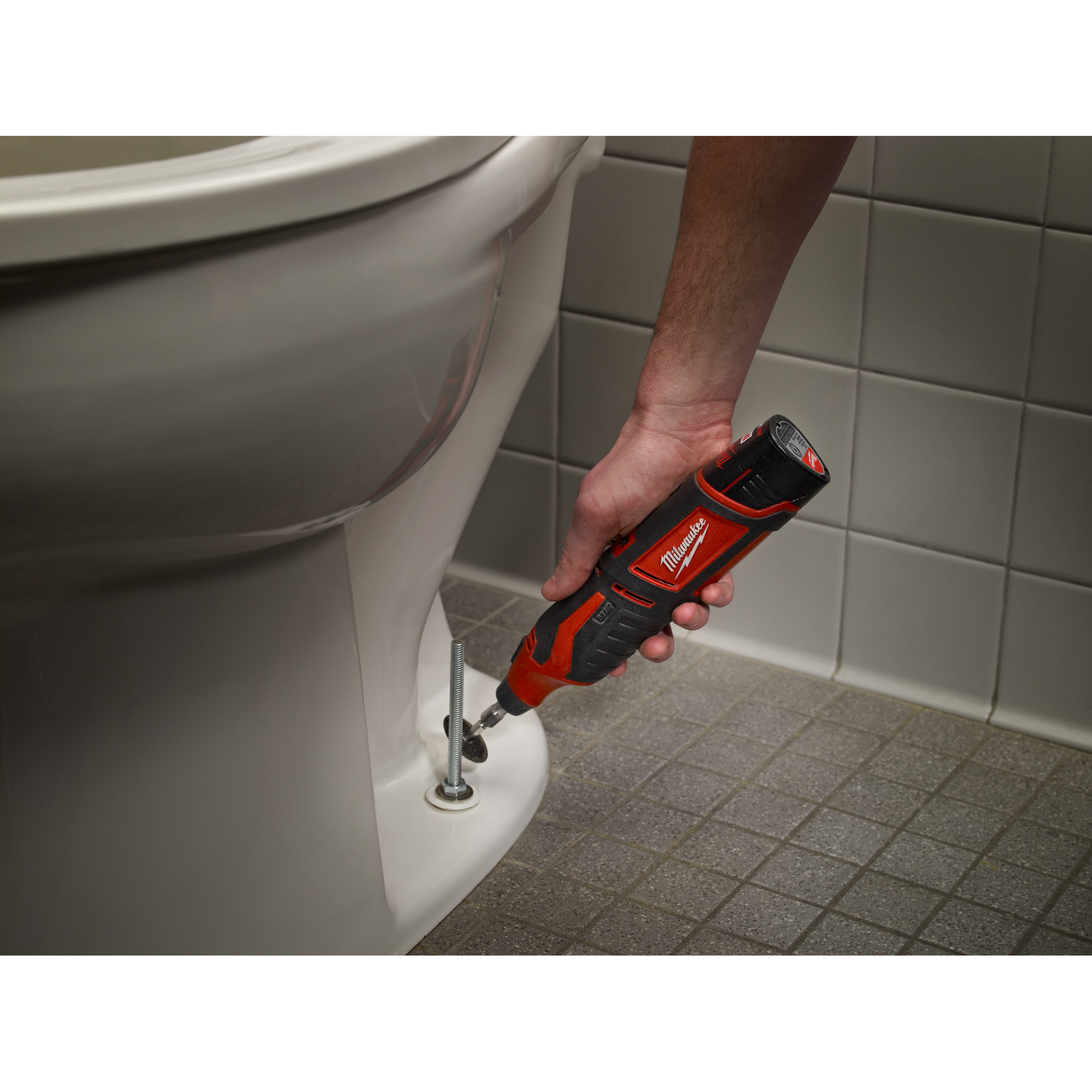 Person using a red and black cordless rotary tool to loosen a bolt on a toilet base for home improvement tasks