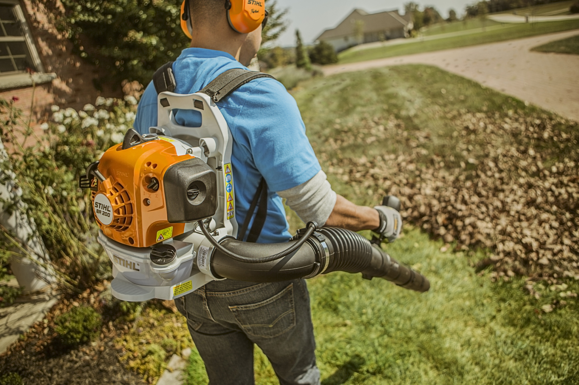 Person using an orange STIHL backpack blower to clear dry leaves from a residential lawn pathway