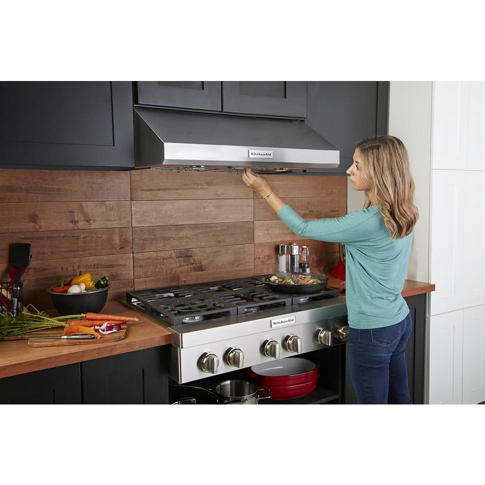 Woman adjusting stainless steel range hood above KitchenAid commercial-style gas cooktop in modern kitchenware collection