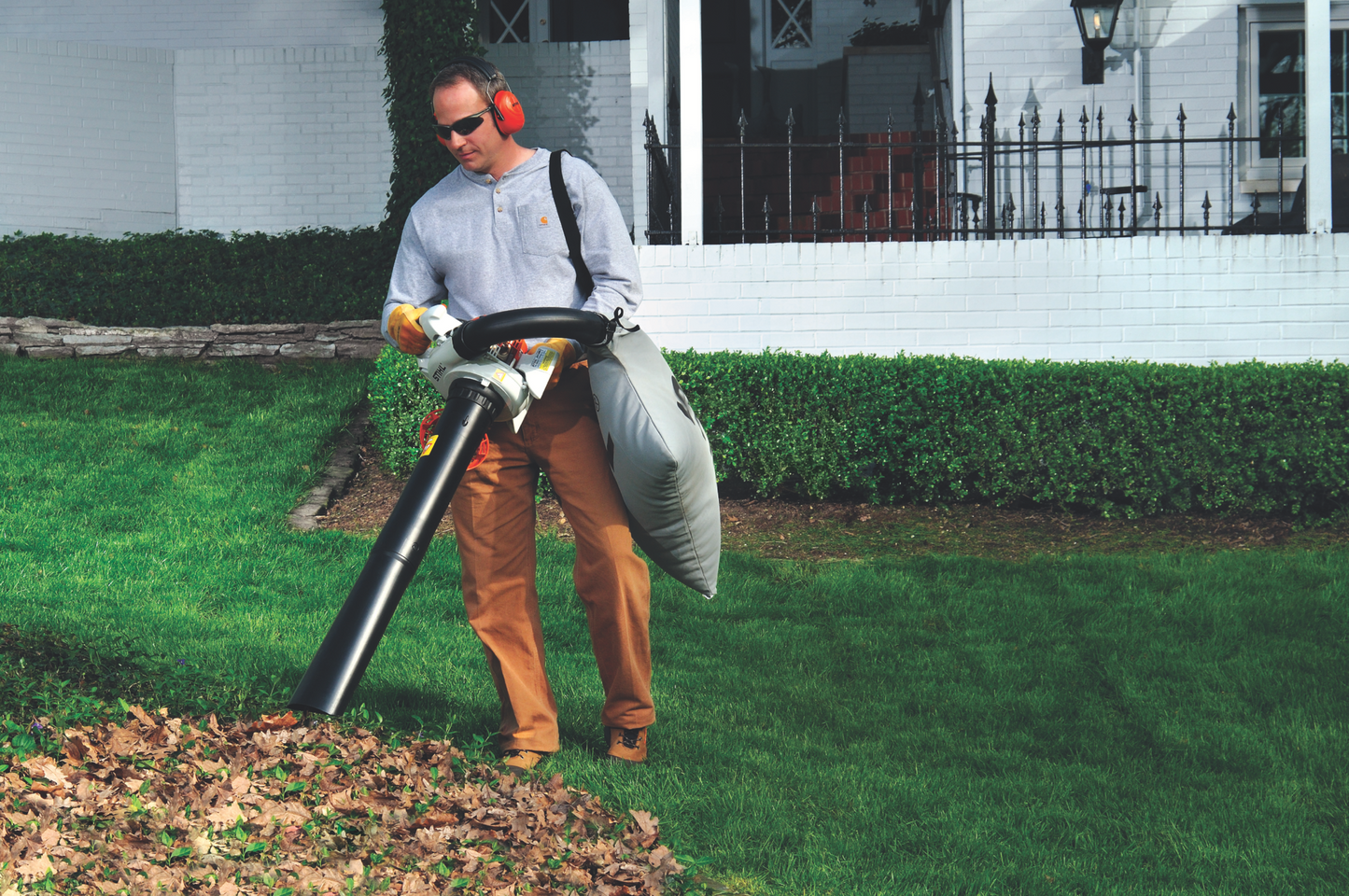 Man wearing ear protection using a gas-powered leaf blower to clear fallen leaves from a lawn near a white house