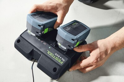 Hands inserting two Festool rechargeable batteries into a dual fast charger for cordless woodworking tools.