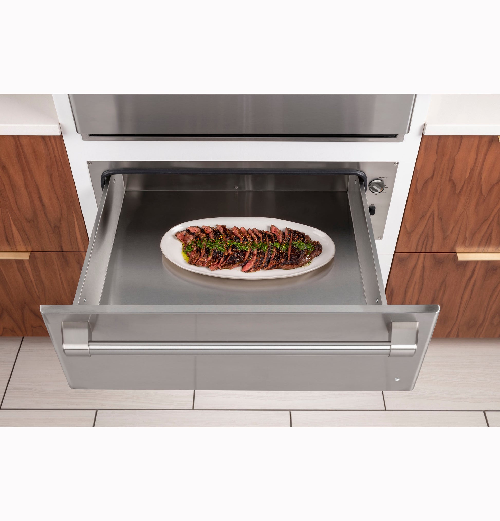 Stainless steel warming drawer open with a plated steak inside, set in modern kitchen cabinetry from appliances collection