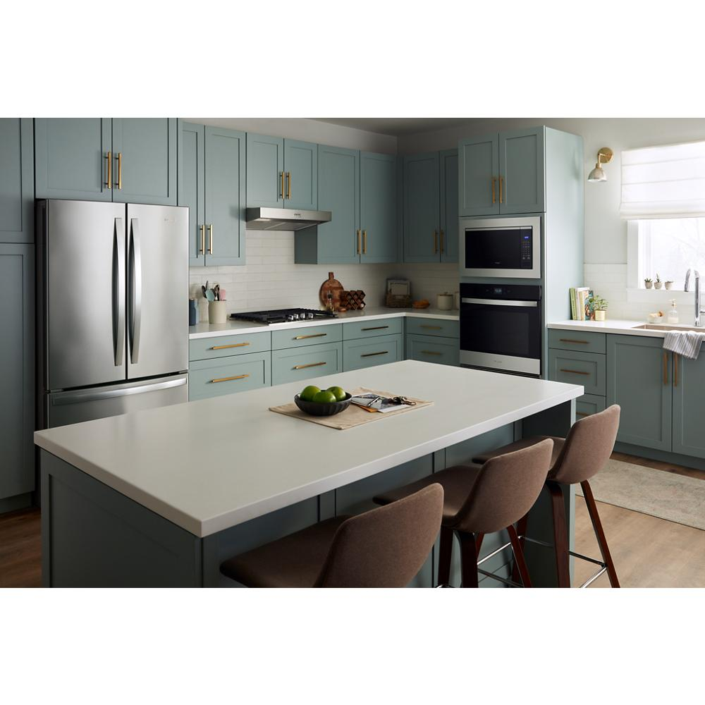 Modern kitchen with muted green cabinetry, gold hardware, stainless steel appliances, and white island with brown chairs