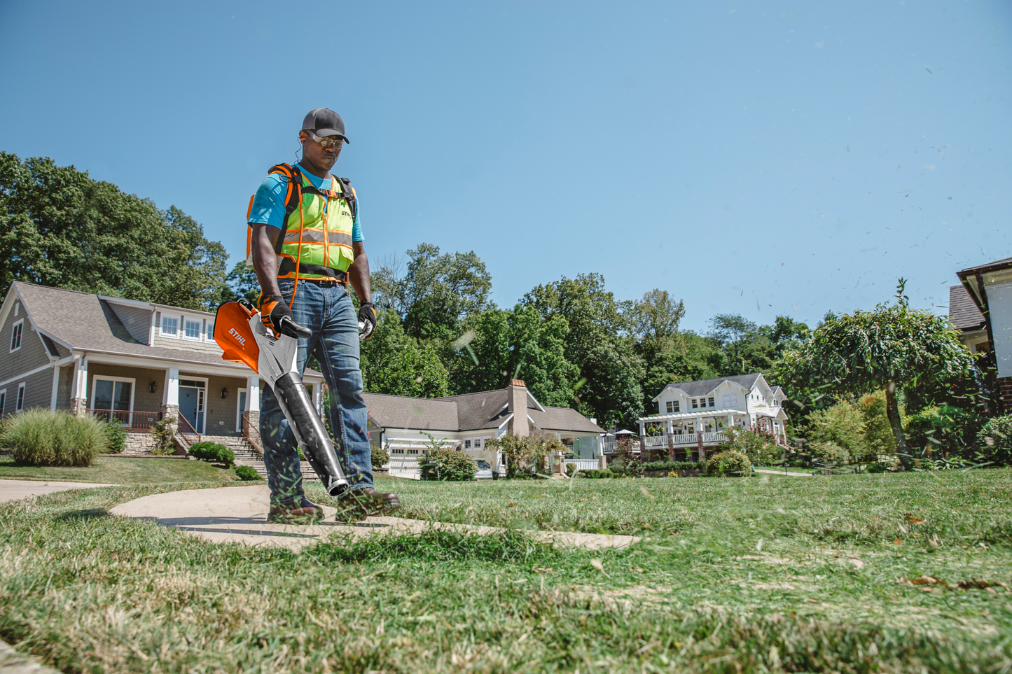 Worker wearing safety vest using orange handheld cordless blower to clear grass clippings from residential lawn edge