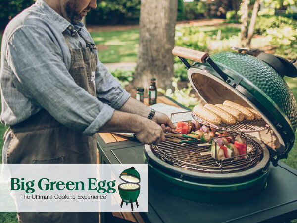 Man wearing an apron grilling corn and vegetable skewers on a green ceramic kamado style grill outdoors in a backyard setting with trees in the background.