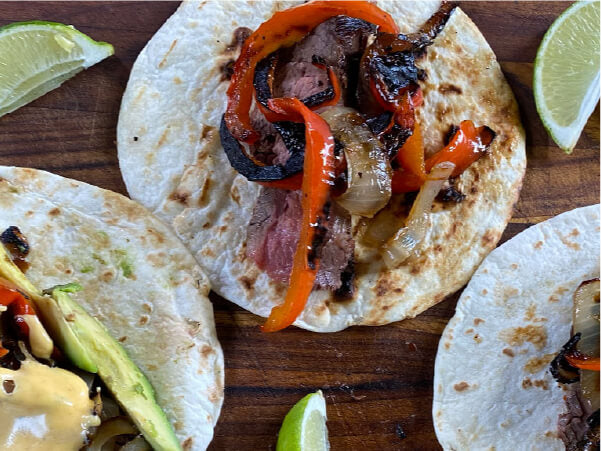 Three soft tortillas topped with grilled strips of beef, charred red peppers and onions, served with lime wedges on a wooden surface.