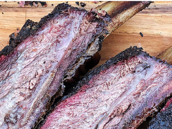 Close-up view of two smoked beef ribs resting on a wooden cutting board, showing textured bark and juicy interior meat.