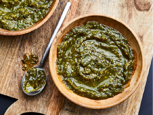 Two wooden bowls filled with thick green salsa on a wooden cutting board, accompanied by a metal spoon with salsa on it.