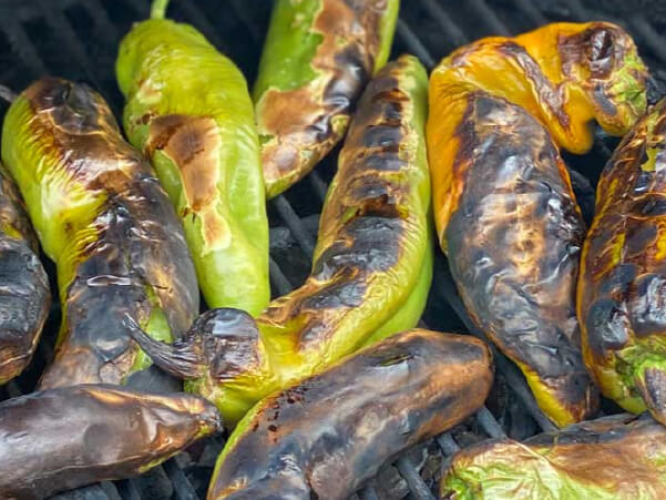 Close-up of several green and yellow peppers roasting on a grill with blackened, charred skins and vibrant colors.