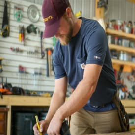 Man wearing a burgundy cap and navy blue shirt focused on woodworking inside a workshop filled with tools and equipment on pegboards.