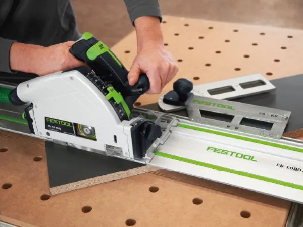 Person using a Festool track saw to cut a black board on a perforated workbench with measuring tools nearby.