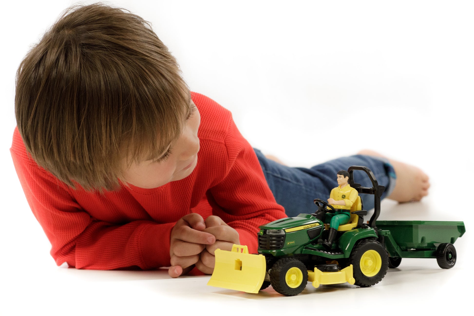 Child in red shirt lying down and playing with green and yellow John Deere tractor toy with trailer and driver figure