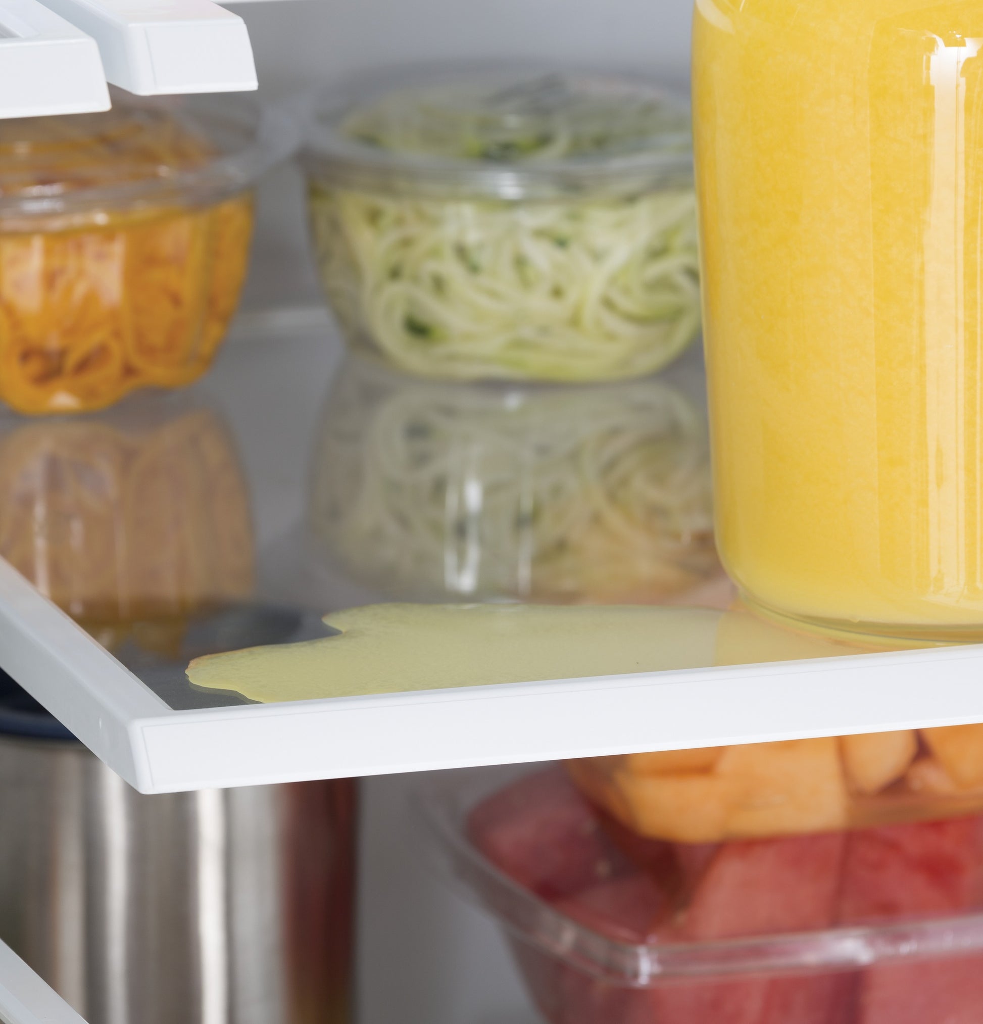 Spilled orange juice pooling on glass shelf inside a refrigerator near containers with fresh food.