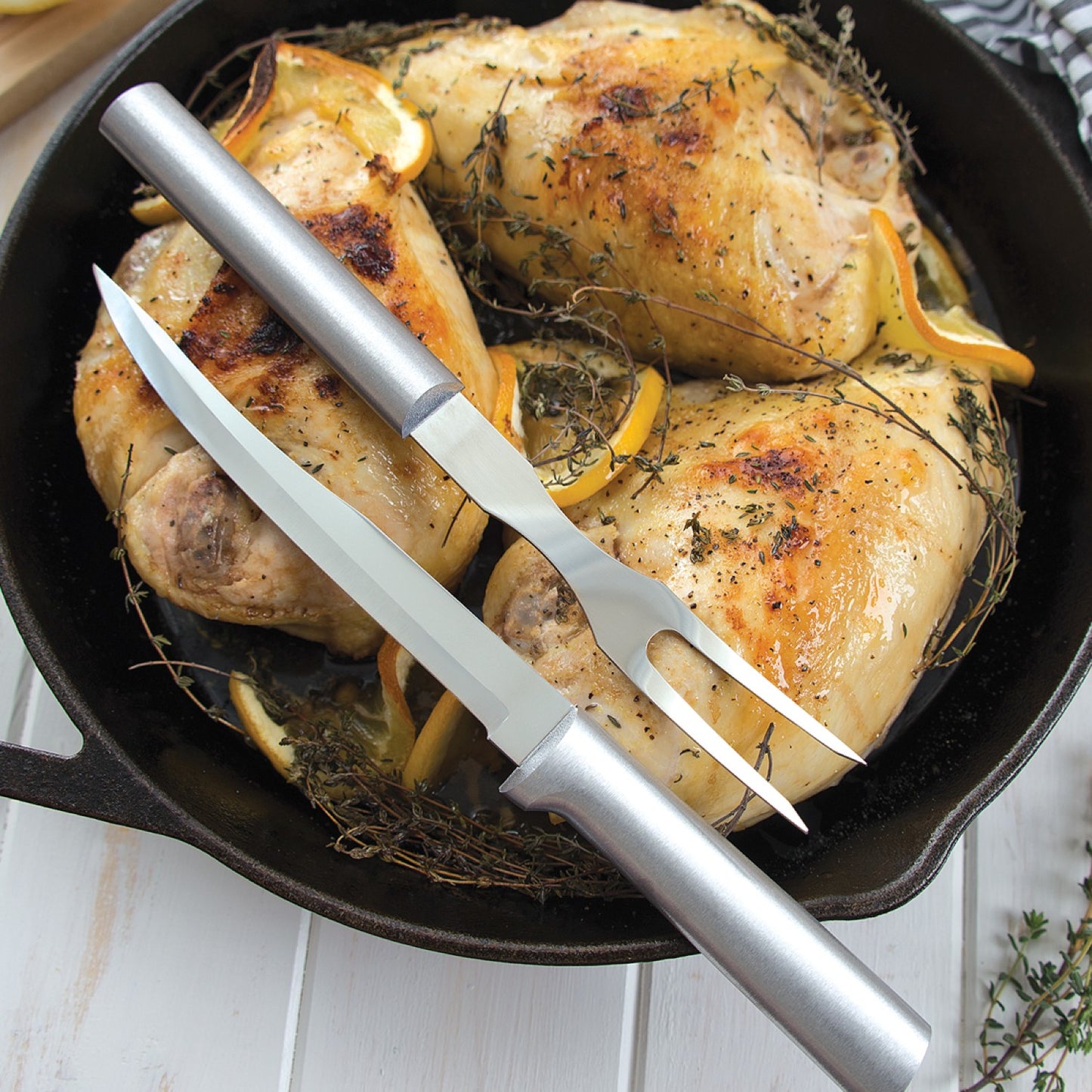 Two stainless steel carving tools with smooth handles resting on cast iron skillet with roasted chicken and herbs from kitchen utensils collection