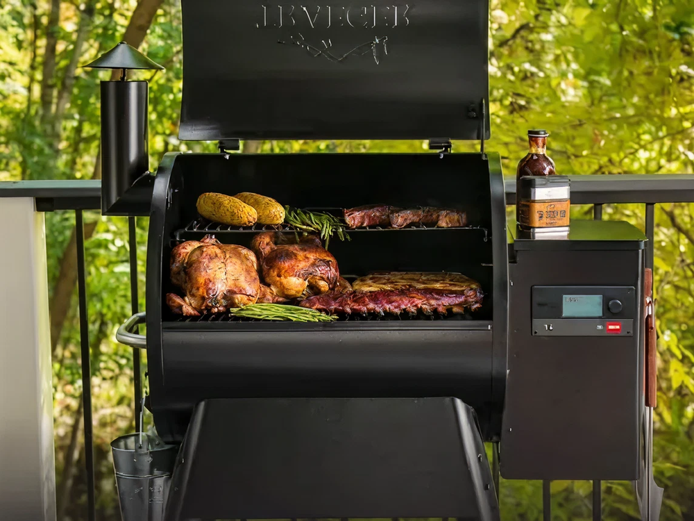 Outdoor black metal smoker grill with open lid showing roasted chickens, corn, ribs, and a bottle placed on the side against a green leafy background