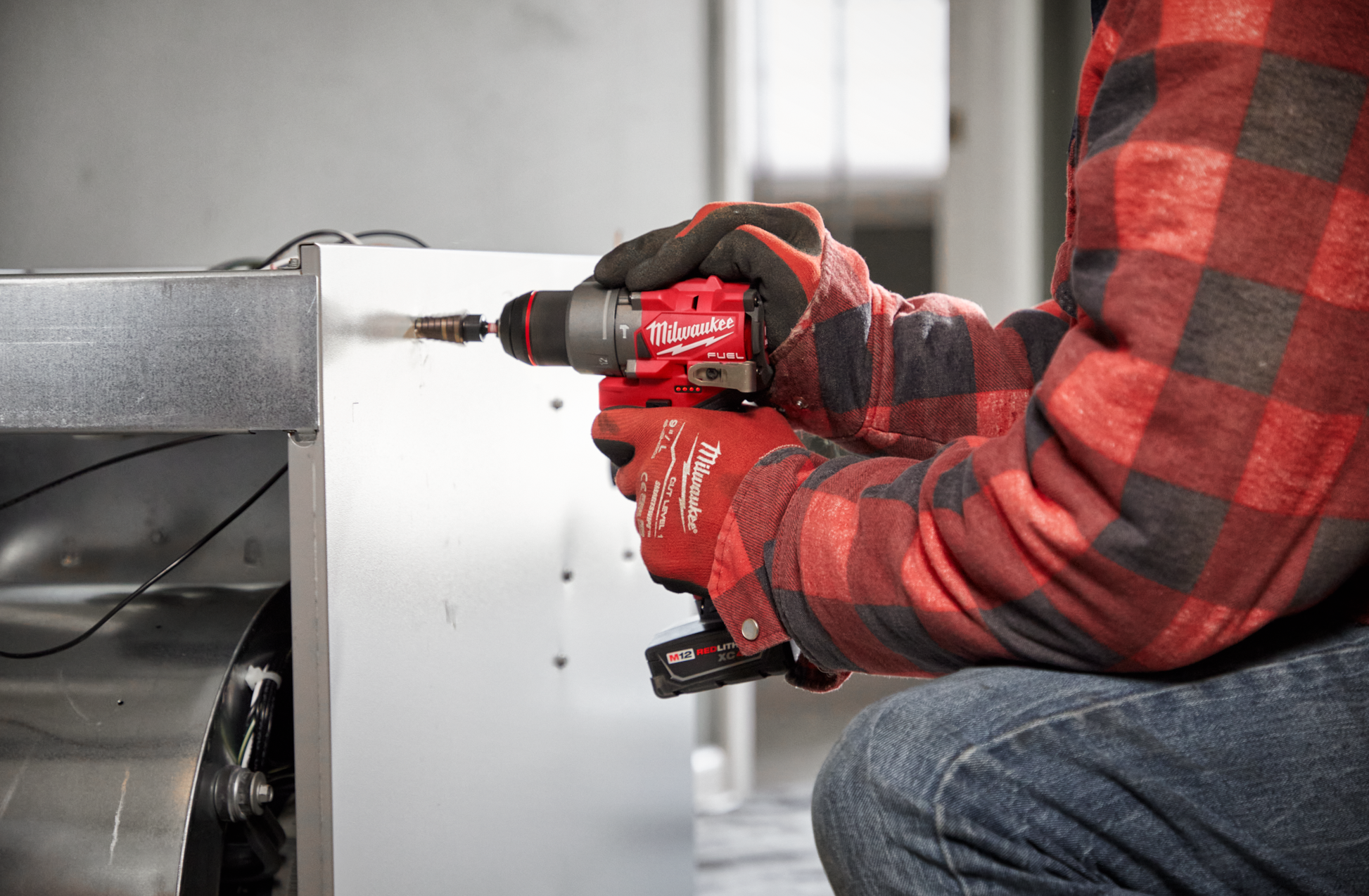 Person wearing gloves and plaid shirt using a red cordless drill driver on metal in a woodworking power tools setting