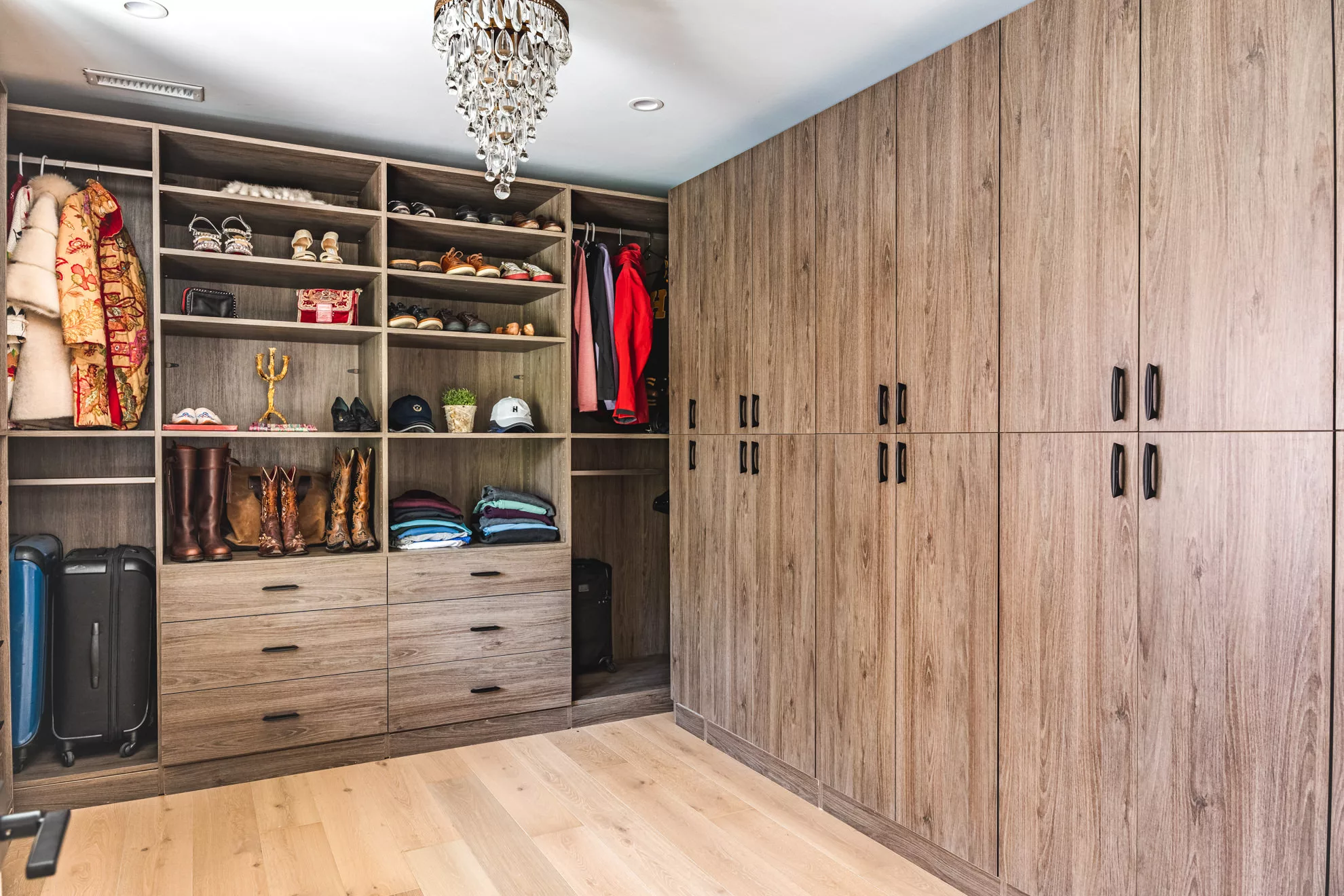 Spacious walk-in closet with light wood cabinetry and shelving holding coats, shoes, folded clothes, luggage, and a crystal chandelier overhead.