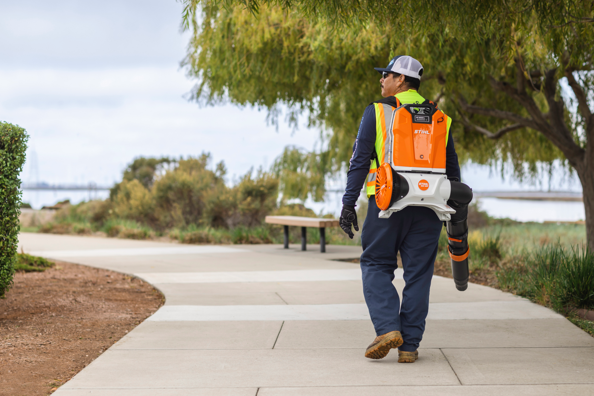 Person carrying backpack-style battery blower with orange and white casing along a paved path outdoors