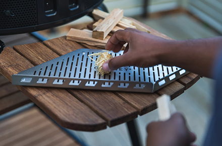 Triangular metal wood rack set on wooden table with hand placing kindling, part of grilling tools collection