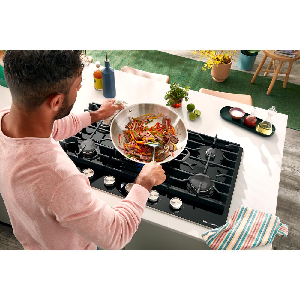 Person stirring vegetables in stainless steel pan on black glass 30" Gas-on-Glass Cooktop in modern kitchen setting
