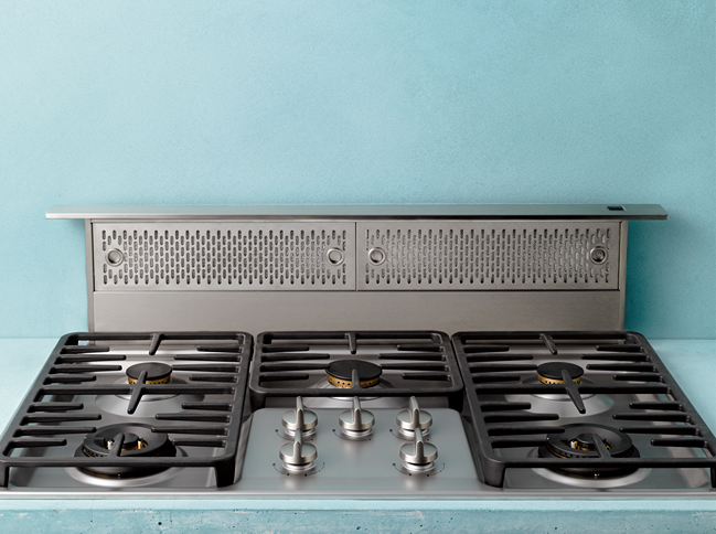 Brushed stainless steel downdraft vent with perforated filters behind gas cooktop burners in kitchen appliance setting