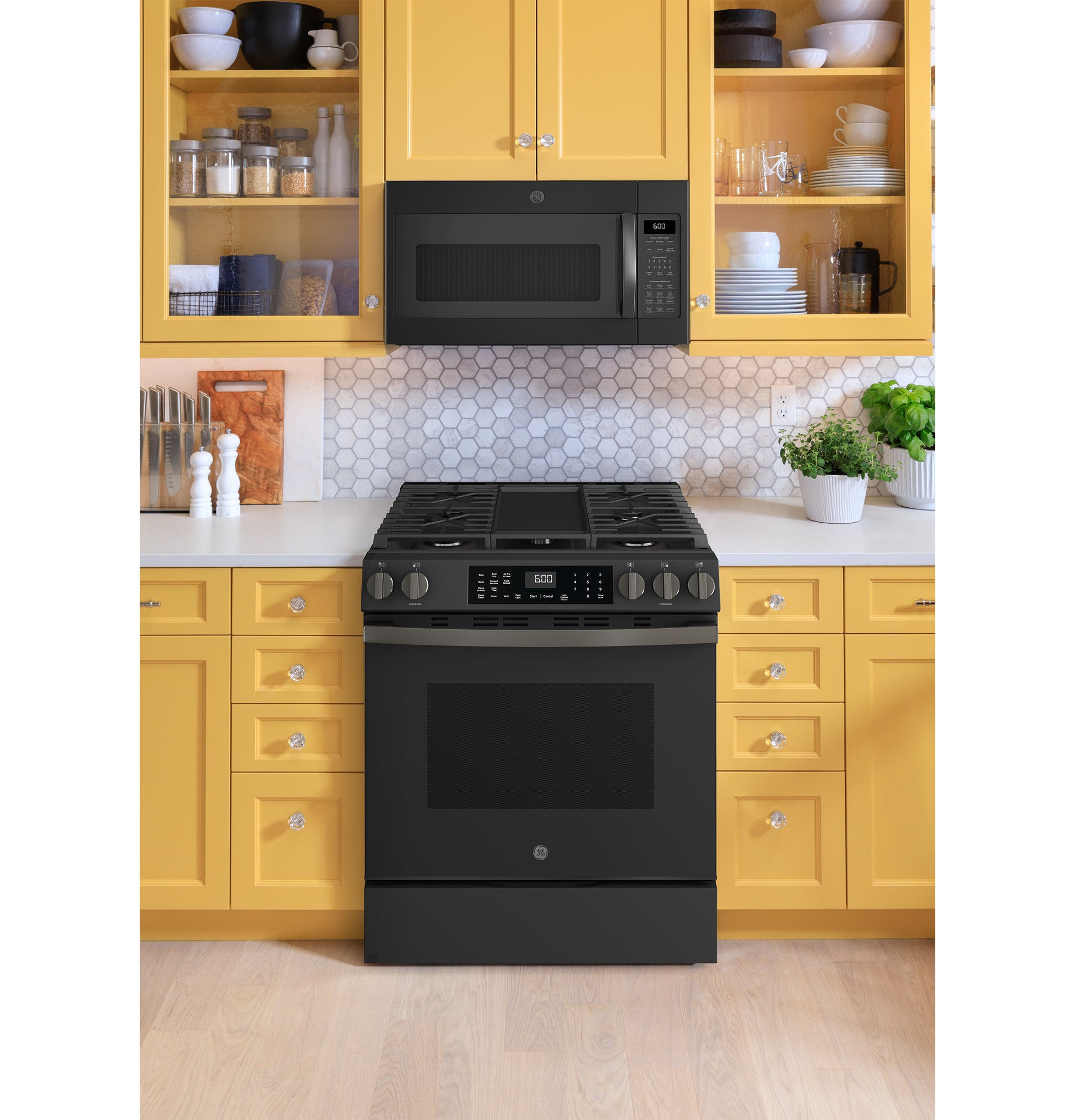 Black slide-in gas range with digital control and microwave mounted above, set in yellow kitchen cabinetry with hex tile backsplash