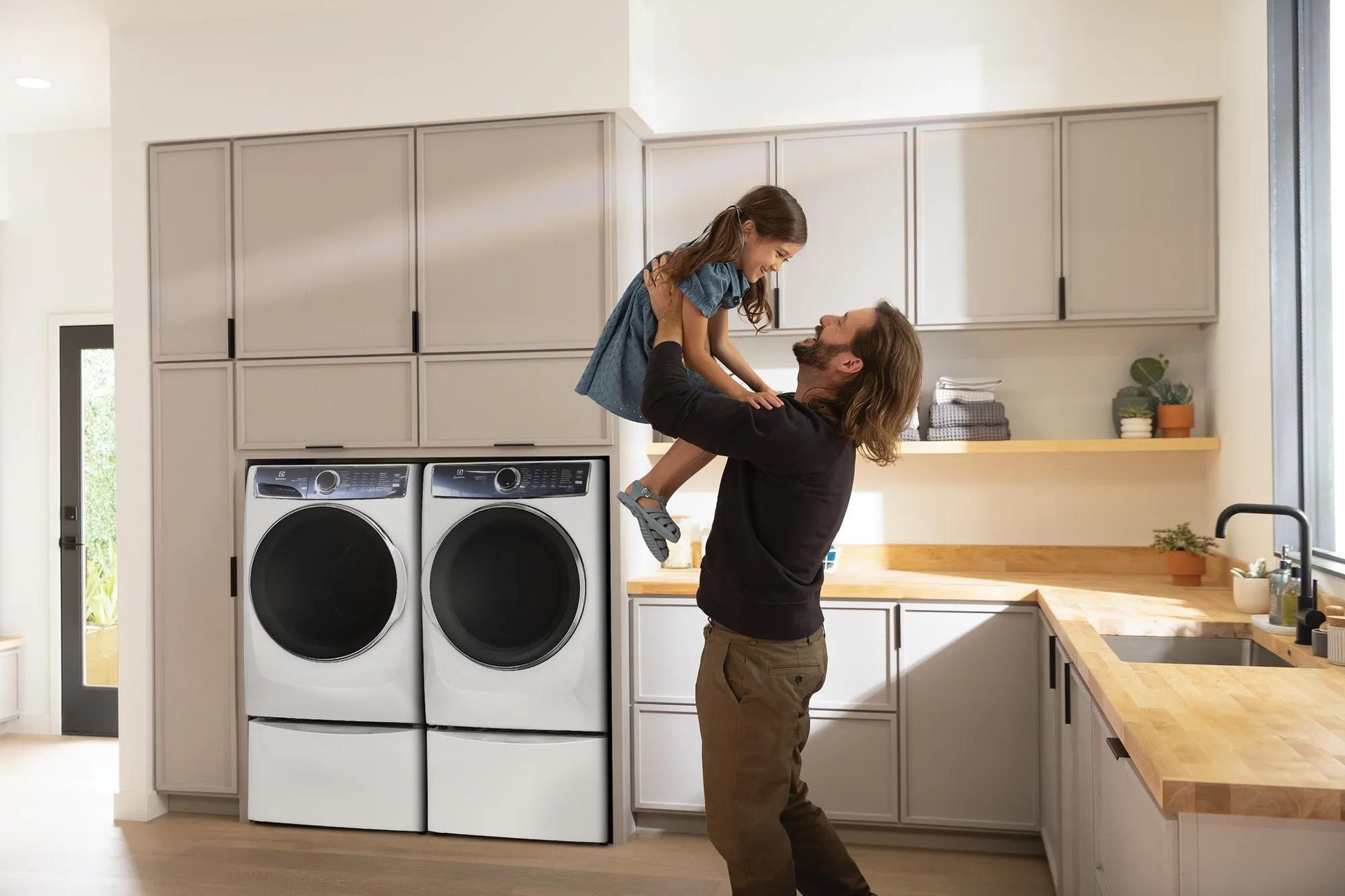 Modern front load washer and dryer set in beige kitchen with man lifting child by wooden countertop in appliances setting