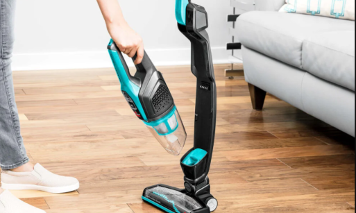 Person detaching electric blue cordless stick vacuum handheld unit from main body on hardwood floor near couch