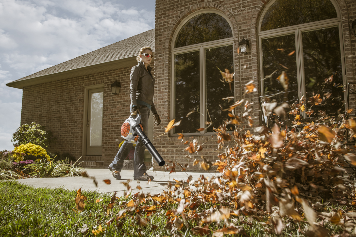 Person using a gas-powered handheld blower to clear leaves from a walkway outside a brick house during home maintenance