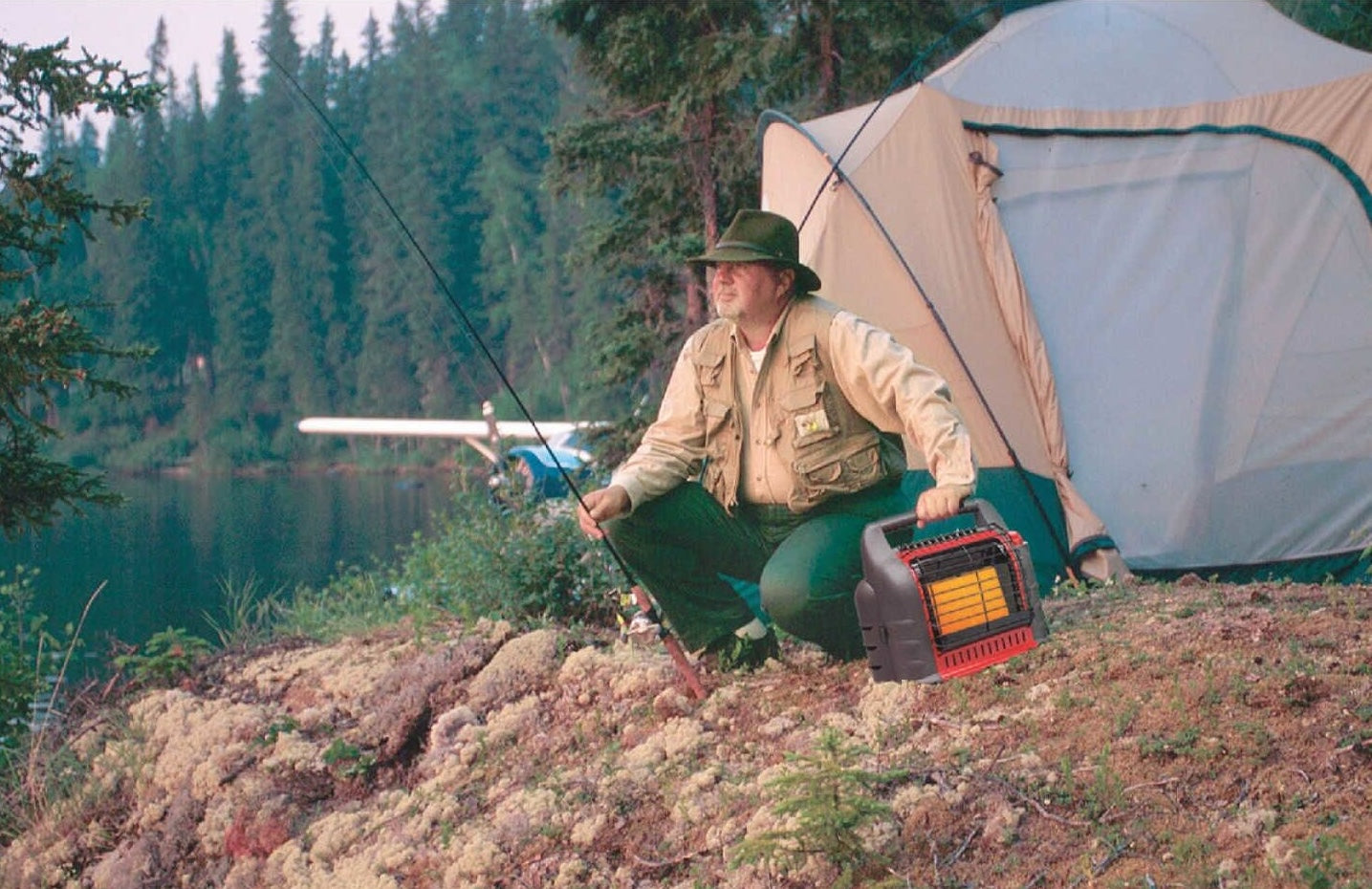 Man fishing by a lake next to a tent holding a portable radiant propane heater in All Products and Home Improvement collections