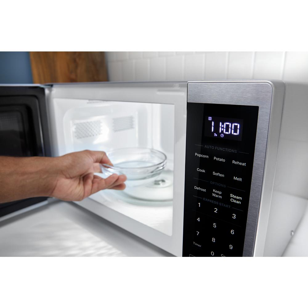 Hand placing glass bowl inside black countertop microwave with digital control panel and LED display in kitchen setting